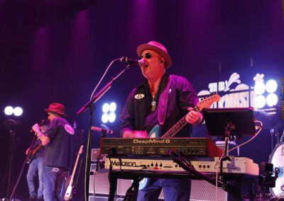 A musician wearing a hat and sunglasses sings and plays electric guitar onstage, with a keyboard in front of him. Another band member is in the background, and colorful stage lights create a vibrant atmosphere.