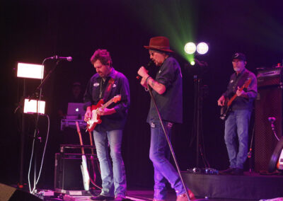A band performs on stage with one person playing electric guitar, another singing into a microphone, and a third playing bass. Stage lights and equipment are visible in the background.