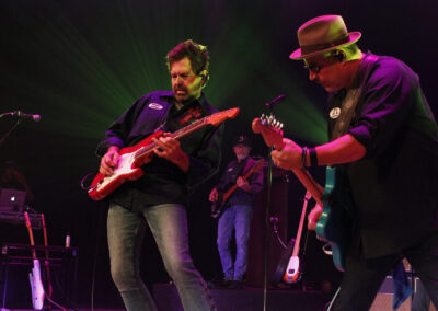 Two guitarists passionately perform on stage under colorful lights, both wearing dark shirts and jeans. Other band members and musical equipment are visible in the background.