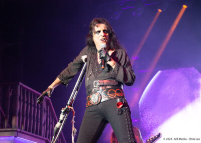 A rock musician with dramatic face makeup and dark clothes sings energetically on stage, gripping a microphone stand. Bright stage lights and a large circular backdrop highlight the performance.