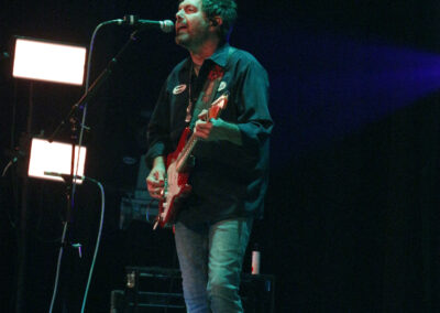 A musician wearing a dark shirt and jeans plays an electric guitar and sings into a microphone on stage, with stage lights and equipment visible in the background.