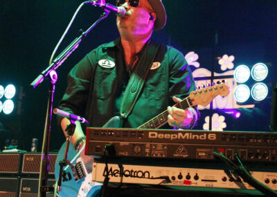 A musician wearing sunglasses and a hat sings into a microphone while playing electric guitar and standing behind multiple keyboards on a stage with colorful lights in the background.