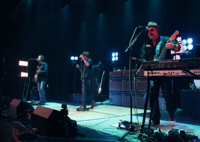 A live band performs on stage under blue lighting. Three musicians play instruments—guitar, keyboard, and harmonica—while singing. Amplifiers and stage equipment are visible in the background.