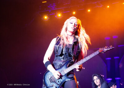 A guitarist with long blonde hair plays an electric guitar on stage under dramatic lighting, with another musician in the background and colorful stage lights above.