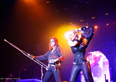 Two musicians perform on stage under dramatic lighting; one holds a long staff, wearing dark clothes and sunglasses, while the other, dressed in black leather, plays an electric guitar raised over her shoulder.