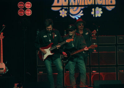 Three musicians perform on stage with electric guitars, standing in front of a wall of VOX amplifiers. The background features a colorful sign, and red stage lights illuminate the scene.