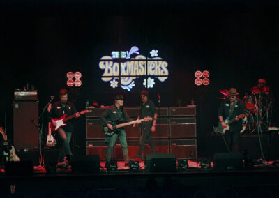 A band performs on stage with guitars and drums under red lighting. The backdrop displays The Boxmasters in stylized text with decorative elements. Five musicians are visible, each focused on their instruments.