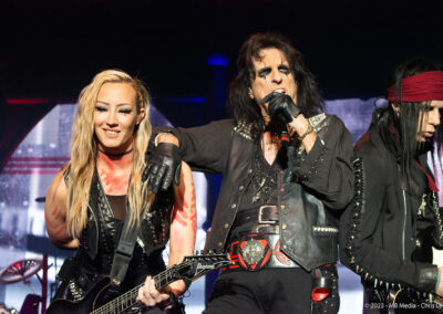 Three rock musicians perform on stage; the woman on the left smiles while playing guitar, the man in the center sings into a microphone with his arm over her shoulder, and another guitarist stands to the right.