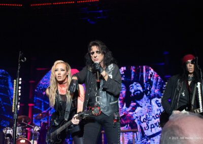Three musicians perform onstage under dramatic lighting. The central figure in black leather holds a cane and sings, while a blonde guitarist and another musician in a bandana play alongside. A colorful, graffiti-like background is visible behind them.