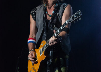 A rock guitarist wearing a hat, sleeveless vest, and patterned pants plays an electric guitar on stage under dramatic lighting.
