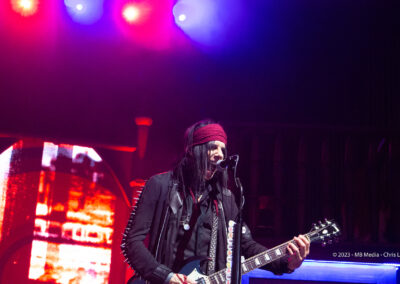 A rock musician with long dark hair, wearing a red headband and black jacket, plays an electric guitar and sings into a microphone on stage under colorful lights.