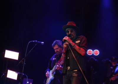 Billy Bob Thornton - A band performs on stage under blue and purple lights; the lead singer in a red hat and sunglasses sings into a microphone, while two guitarists play in the background.