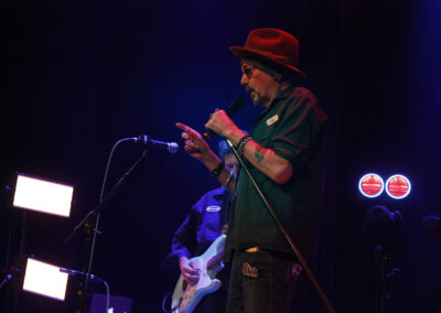 A man wearing a red hat and sunglasses sings into a microphone on stage, gesturing with his hand. Stage lights and a guitarist are visible in the background, creating a vibrant concert atmosphere.