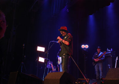 A singer wearing a hat and sunglasses performs passionately on stage with a microphone. Several musicians play instruments in the background under purple stage lights.