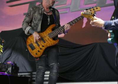 A musician wearing a hat and jacket plays an electric bass guitar on stage, with a large band logo displayed on a screen behind him. Another performer is partially visible on the right.