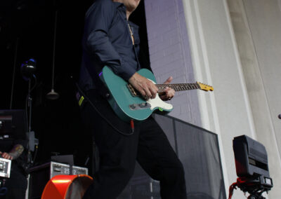 A musician wearing a black hat and dark clothing plays an aqua electric guitar on stage, with stage equipment and a white wall in the background.