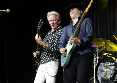 Two guitarists perform energetically on stage, one wearing a patterned shirt and white pants, the other in a black hat and dark shirt. A drum set with a visible band logo is in the background.