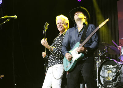 Two guitarists perform energetically on stage under bright lights; one smiles at the audience while the other, wearing a hat, focuses on playing. A drum set with Felder on the bass drum is visible in the background.