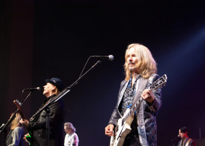A rock band performs on stage with multiple members playing guitars and singing into microphones, illuminated by bright stage lights against a dark background.