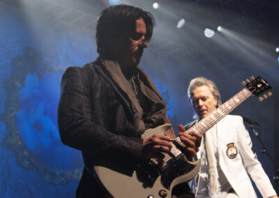 Two musicians perform on stage; one in the foreground plays an electric guitar while wearing dark clothing, and the other stands behind in a white outfit. Stage lights and a decorative backdrop are visible.