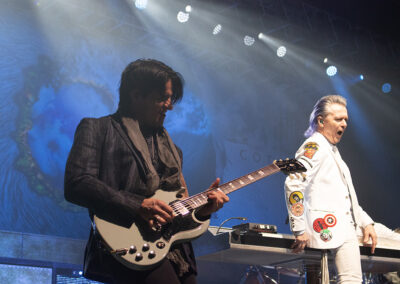 Two musicians perform on stage. The guitarist on the left plays energetically, while the keyboardist on the right, dressed in a white suit with colorful patches, sings passionately. Stage lights and a colorful backdrop create a dramatic scene.
