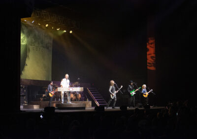 A rock band performs on stage with dramatic lighting, featuring a keyboardist at center, guitarists, and a drummer, in front of an audience in a dark concert venue.