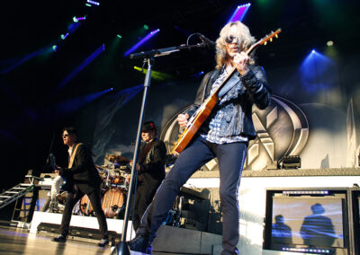 A rock band performs on stage with dramatic lighting; the guitarist in front wears sunglasses and a leather jacket, while other band members play in the background. A large circular band logo is displayed behind them.