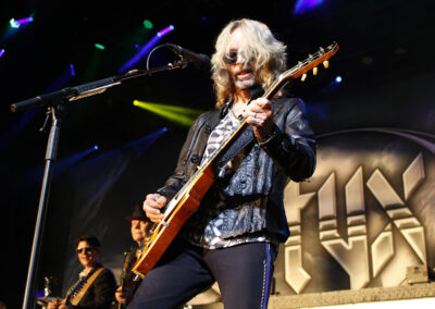 A guitarist with long blond hair and sunglasses plays an electric guitar on stage, with other band members and a large STYX logo visible in the background under colorful stage lights.