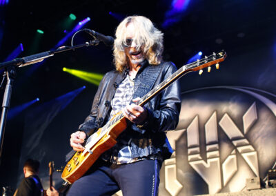 A long-haired musician wearing sunglasses plays an electric guitar on stage, with colorful lights above and a large logo reading STYX in the background.