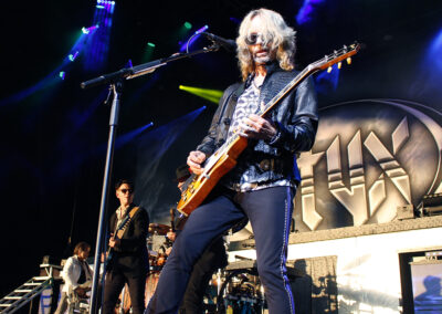 A musician with long blond hair and sunglasses plays an electric guitar on stage with a live band. Colorful stage lights shine above, and a large STYX sign is visible in the background.