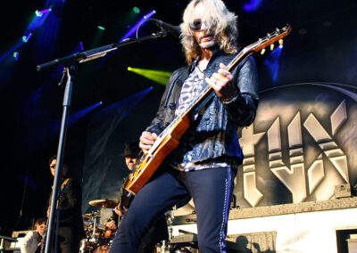 A musician with long blonde hair and sunglasses plays an electric guitar on stage, with band members and drums in the background. A large STYX band logo is displayed behind them under colorful stage lights.