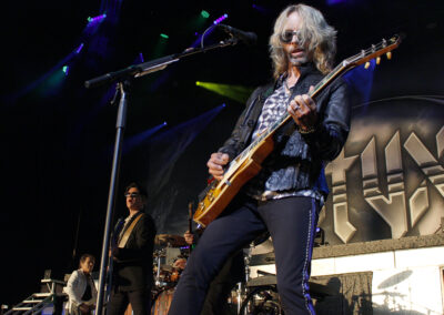 A rock band performs on stage with colorful lights; the guitarist in sunglasses stands front and center, playing energetically while other band members play instruments in the background. The bands logo is visible behind them.