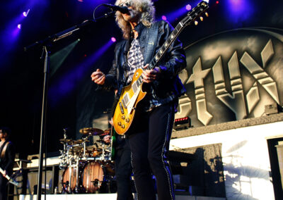 A musician playing an electric guitar sings into a microphone on stage, with drums and large STYX logo in the background, under colorful stage lights.