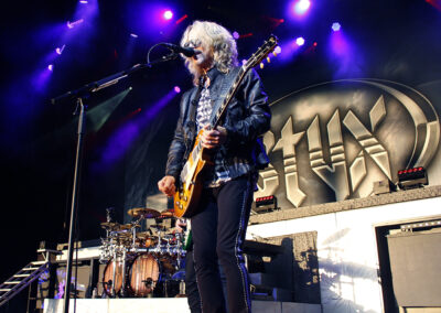 A guitarist with long gray hair plays an electric guitar and sings on stage with purple lighting and a large STYX band logo in the background; a drummer is visible behind him.