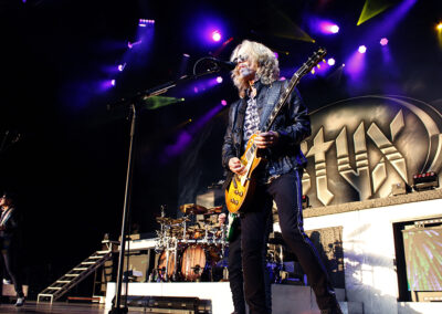 A guitarist with long hair performs on stage under colorful lights, standing in front of a drum set and a large STYX band logo backdrop. Other band members and instruments are visible in the background.