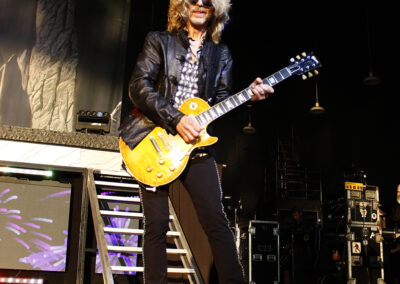 A musician with long blonde hair and sunglasses plays an electric guitar on stage. He wears a black leather jacket and dark pants. Stage equipment, lighting, and a staircase are visible in the background.