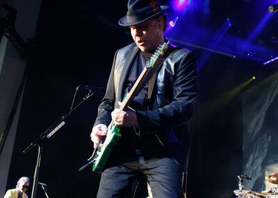 A guitarist in a black suit and hat plays an electric guitar on stage under purple and blue lights, with other musicians performing in the background.