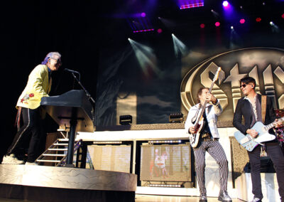 Three musicians perform energetically on stage with guitars and a keyboard, under colorful stage lights. A large STYX logo is visible in the background. The atmosphere suggests a lively rock concert.