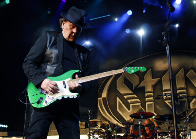 A musician in a black outfit and hat plays a bright green electric guitar on stage, with colorful lights above and a drum set and large Styx logo visible in the background.
