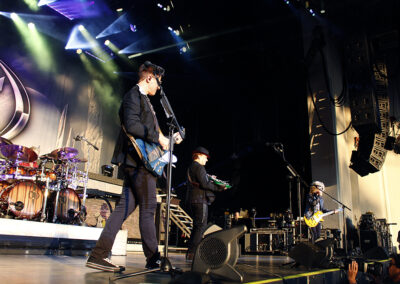 A rock band performs live on stage with colorful lights above. Three musicians play guitars and sing, while a drummer is visible on the left. Large speakers and various instruments are set up around them.