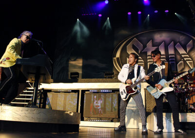A rock band performs on stage with colorful lighting; the keyboardist is on the left in a yellow jacket, while two guitarists and a drummer play in front of a large Styx logo in the background.