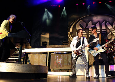 Three musicians perform energetically on stage under colorful lights, with one on keyboard and two playing guitars. The large backdrop features the word Styx in bold letters. The scene is lively and dynamic.