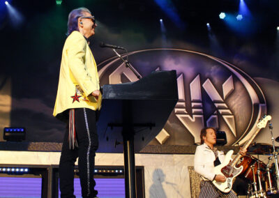A musician in a yellow jacket stands at a keyboard on stage, while another musician plays guitar nearby. Colorful stage lights and a large STYX band logo are visible in the background.