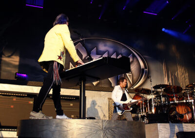 Two musicians perform on stage under purple lights; one plays a keyboard while wearing a yellow jacket, and the other plays a guitar in a white jacket next to a drum set. A large logo is visible in the background.