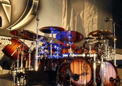 A large, elaborate drum kit with multiple drums, cymbals, and stands is set up on a stage with dramatic lighting and a textured backdrop. The drum shells are a rich wood finish with shiny metal hardware.