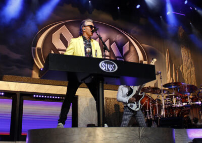 A musician in a yellow jacket plays a keyboard on stage with the Styx logo, dramatic lighting, and bandmates playing instruments in the background during a live concert.