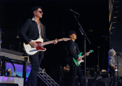 Three musicians perform on stage with electric guitars. The musician in front wears sunglasses and a dark suit, another behind him wears a black hat, and a third stands further back, illuminated by stage lights.