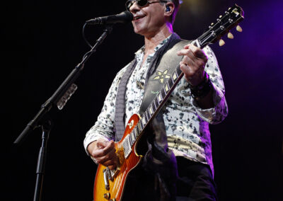 A musician wearing sunglasses and a patterned shirt plays an electric guitar and sings into a microphone on stage under purple lighting.