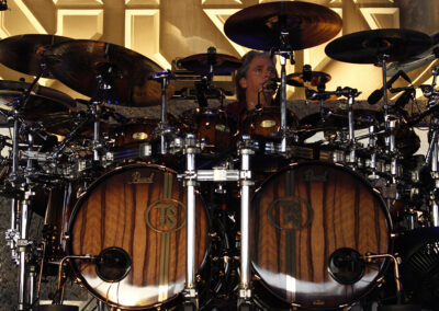 A drummer surrounded by an elaborate drum kit with multiple cymbals and wooden bass drums, performing on stage with a lit background.