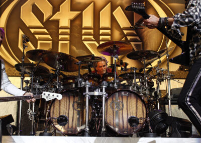 A drummer plays an elaborate drum set onstage, flanked by two guitarists. Behind them is a large gold and white STYX band logo as part of the backdrop. The stage is brightly lit.